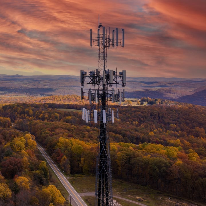 Aerial view of a tall cellular communications tower with multiple antenna arrays, rising above a forested landscape with autumn foliage in orange, yellow, and green, a winding two-lane road below, and a dramatic orange and pink sunset sky