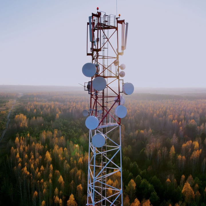 Aerial view of a red-and-white telecommunications tower with multiple microwave dishes and antennas, rising above a misty forest of green and autumn-gold trees