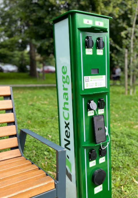 Green NexCharge charging station beside a park bench, with a smartphone plugged in to charge