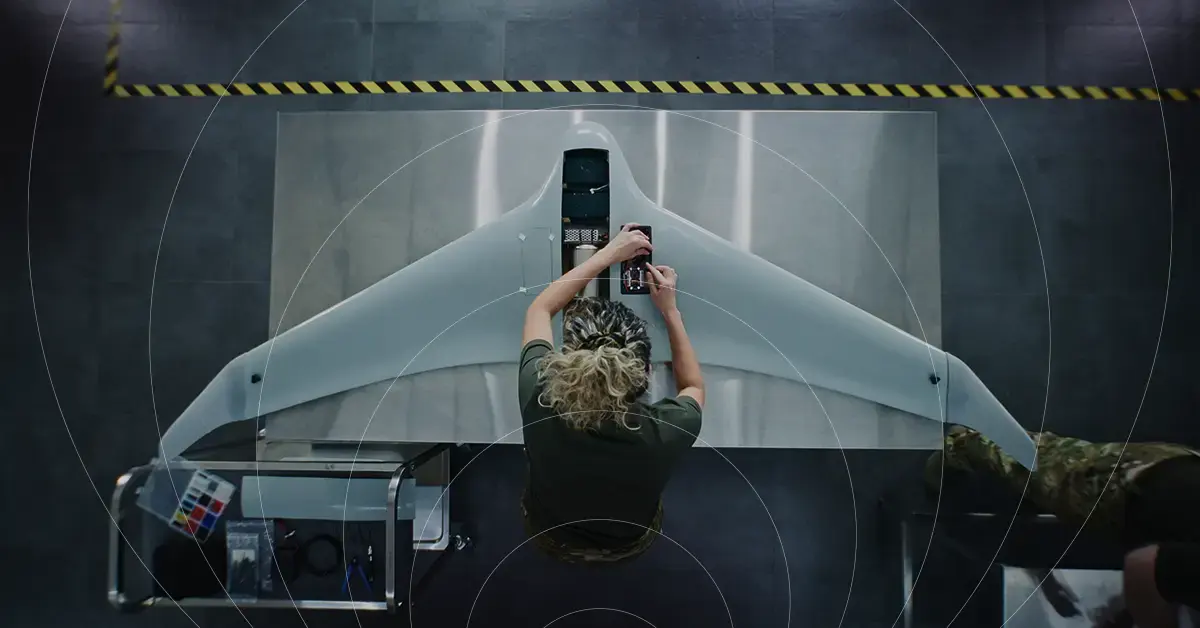 Overhead view of a technician configuring a military drone's onboard IoT sensors, with icons for drone connectivity, AR/smart-goggle interface, and ground-vehicle telemetry alongside.