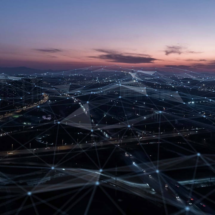 Et skumringsbilde fra luften av et motorveikryss i byen med lysende trafikklys, overlagret med et gjennomskinnelig nettverk av sammenkoblede noder og linjer som symboliserer global IoT-tilkobling.