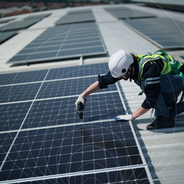 Engineer in a white hard hat and high-visibility vest kneeling on a rooftop solar array, inspecting a panel, with a faint circle graphic overlay.