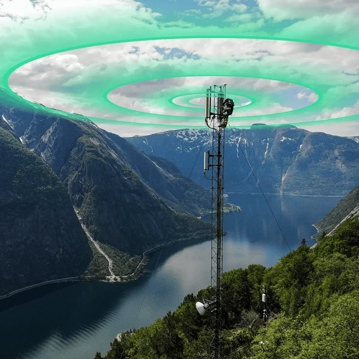 Cellular network tower on a mountain ridge overlooking a Norwegian fjord, with a technician working at the top and green signal rings radiating outward to represent network coverage.