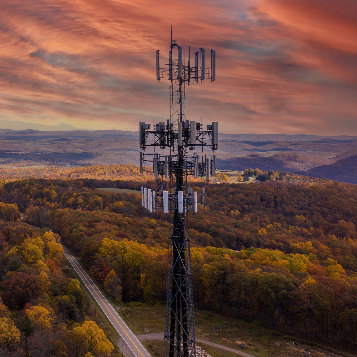 Aerial view of a tall cellular communications tower with multiple antenna arrays, rising above a forested landscape with autumn foliage in orange, yellow, and green, a winding two-lane road below, and a dramatic orange and pink sunset sky