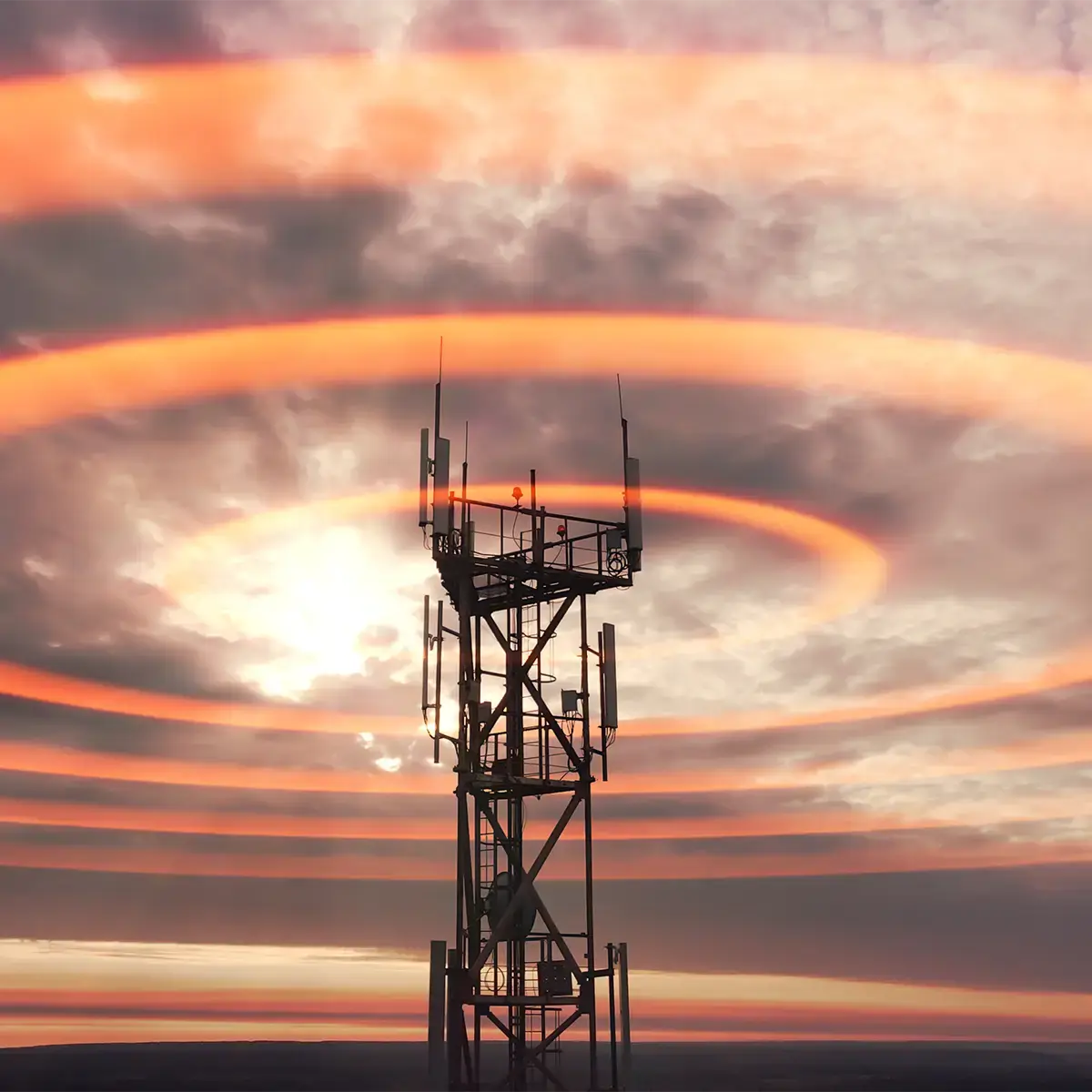 A cellular network tower silhouetted against a dramatic orange sunset sky, with concentric ripple-like rings radiating outward from the antennas across the clouds to suggest wireless signal propagation.